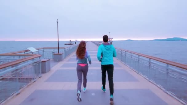 Ung hälsosamt liv par kvinna man jogga på piren stranden tidig morgon moln atletisk livsstil himmel blå Uhd — Stockvideo
