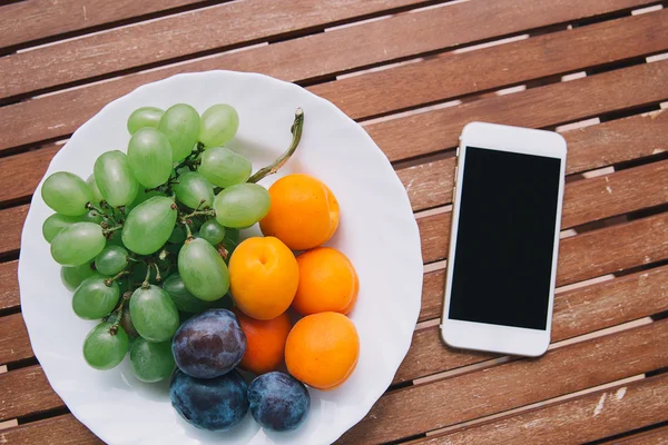 Placa con frutas frescas mezcladas en el fondo de madera con teléfono. Surtido de frutas jugosas en mesa de madera — Foto de Stock