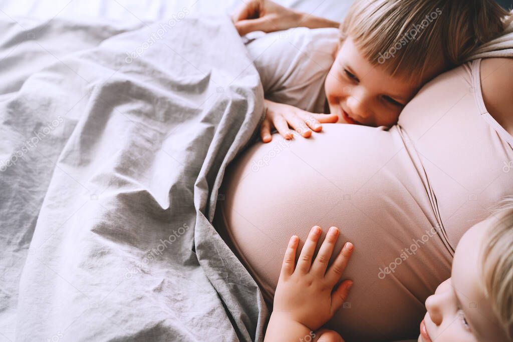 Little kids hold hands on pregnant belly of mom. Pregnant woman with her children relaxing in bed. Loving mother and toddlers together at home. Third pregnancy. Maternity, family, parenting concept.