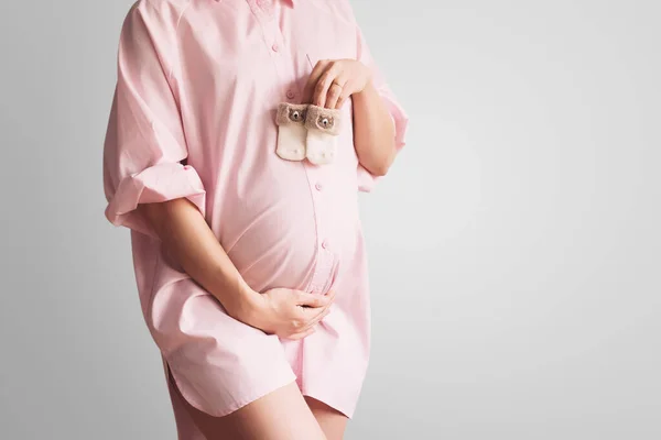 用可爱的小婴儿袜盖住怀孕的肚子 怀孕的概念 漂亮的孕妇穿着粉色衬衫 背景是白色的 年轻母亲在等孩子出生 — 图库照片