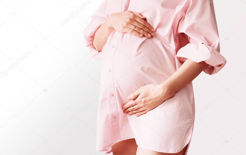 Closeup of beautiful pregnant woman in pink shirt dress holds hands on her belly in white background. Loving mother waiting for baby birth. Pregnancy, maternity, preparation and expectation concept.