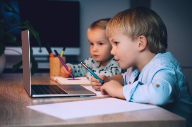 Okul çocuğu ve anaokulu kızı akşam evde, masada dizüstü bilgisayarlarıyla. Dijital teknolojiyi ve internet iletişimini kullanan çocuklar. Çocuklar için çevrimiçi eğitim veya İnternet eğlencesi.