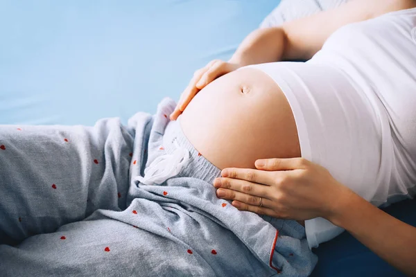 Close-up of beautiful pregnant belly of woman lying in bed. Expectant mother hugging her belly and resting. Loving mother waiting of a baby. Concept of pregnancy, maternity, healthcare, gynecology.