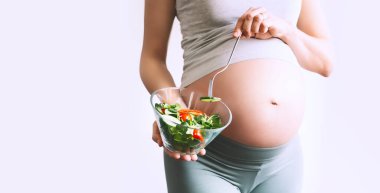 Folik asit içeren sağlıklı yiyecekler yiyen hamile bir kadın, B9 vitamini. Yakın plan hamile kadın göbeği ve sebze salatası. Doğum öncesi beslenme, sağlıklı yaşam tarzı ve hamilelik süresince diyet kavramı.