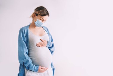 Tıbbi maskeli genç hamile bir kadın göbeğine sarılıyor. Virüsler sırasında konsept koruma ve önleme, ona anne bakımı ve doğmamış bebek sağlığı. Bulaşıcı hastalıklar sırasında hamilelik ve doğum.