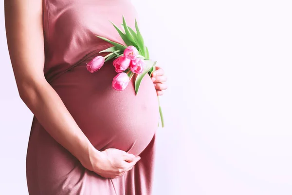 チューリップの花を持つ美しい妊婦は白い背景で腹に手を握っています 産後のピンクのドレスを着た若い女性が出産を待っています 母の日 Holiday Concept — ストック写真