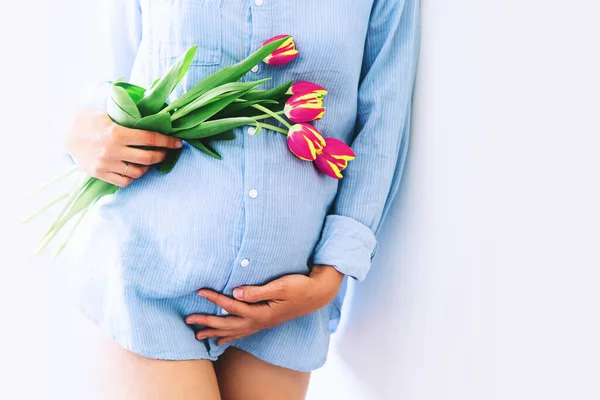 Young pregnant woman with tulips flowers holds hands on belly on a white background. Loving woman in maternity pajamas shirt waiting for a baby birth. Pregnancy, Mother\'s Day Holiday concept.