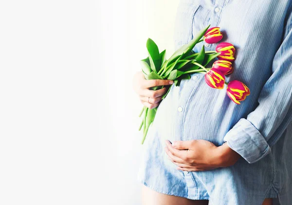 Young pregnant woman with tulips flowers holds hands on belly on a white background. Loving woman in maternity pajamas shirt waiting for a baby birth. Pregnancy, Mother\'s Day Holiday concept.