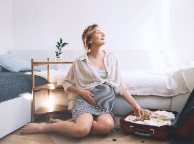 Hamile kadın doğum hastanesi için bavul hazırlıyor. Doğum için hazırlanıyor. Evinde bebek kıyafetleri taşıyan mutlu bir anne. Hamilelik süresince yeni doğan için hazırlık.