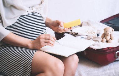 Hamile kadın kontrol listesiyle birlikte hastane çantalarını topluyor. Bebek kıyafetleri ve ihtiyaçlarıyla dolu bir valiz taşıyacak hamile bir anne. Yeni doğuma, doğuma, doğuma hazırlanıyor. Kağıt günlüğe notlar yazıyor..