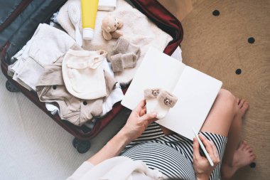 Hamile kadın kontrol listesiyle birlikte hastane çantalarını topluyor. Bebek kıyafetleri ve ihtiyaçlarıyla dolu bir valiz taşıyacak hamile bir anne. Yeni doğuma, doğuma, doğuma hazırlanıyor. Kağıt günlüğe notlar yazıyor..