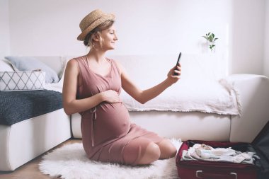 Hamile kadın hastane ya da yolculuk için bavul hazırlıyor, yeni doğan bebek, doğum, doğum, liste yapmak ya da akıllı telefonla fotoğraf çekmek için hazırlanıyor. Temiz kıyafetler ve çantadaki eşyalar. Hamilelik sırasında seyahat.