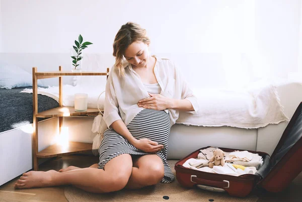 Pregnant woman is packing suitcase for maternity hospital getting ready for childbirth. Happy young mother with travel luggage of baby clothes at home. Preparation for newborn birth during pregnancy.