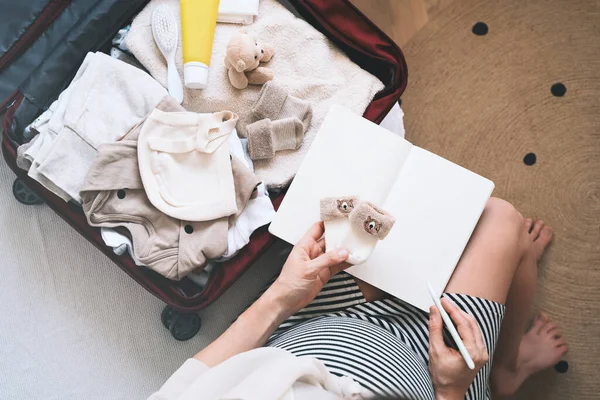 Pregnant woman packing hospital bag with checklist. Expectant mother with travel suitcase of baby clothes and necessities preparing for newborn birth, labor, making notes in paper diary.