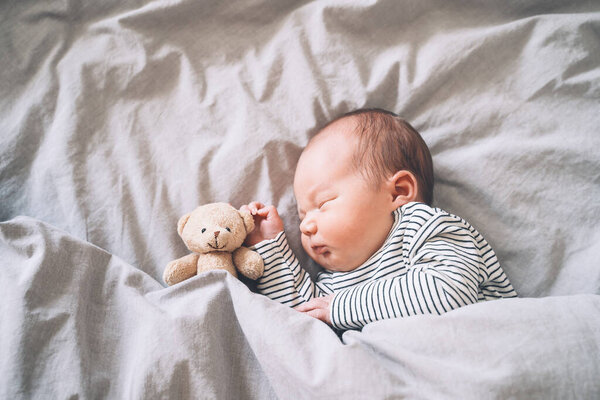 Сон новорожденного ребенка в первые дни жизни. Портрет новорожденного мальчика недельной давности, мирно спящего с милой мягкой игрушкой в кроватке на тканном фоне.