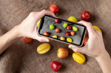 İki el, üzerinde fotoğraf olan bir telefon ve çuval bezinin üzerinde duran karambola elmaları tutuyor. Yemek Fotoğrafçısı Telefonda Meyve Çekiyor