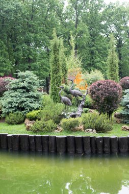 Parkta peyzaj, bir sürü farklı bitki, bir göl ve bir leylek heykeli.