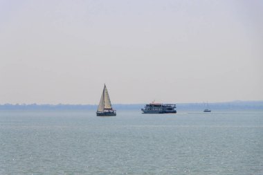 BALATONFURED, HUNGARY. Macaristan, Balaton Gölü 'ndeki yat ve turistik gemi zümrüt renginde berrak sularda yüzüyor ve Balatonfured kasabası kıyı şeridinde görülebiliyor..