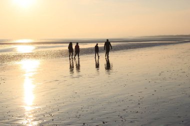 Gün batımı siluetinde düz kumsalda yürüyen 4 kişilik bir aile ve düşük gelgit arka planında fotokopi alanı ile ıslak kumu yansıtan bir aile.