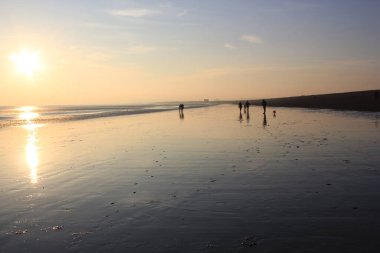 Günbatımında yürüyen insanlar, Winchelsea plajı ile Rye Harbour doğası arasındaki kumsaldaki ıslak kumu yansıtırken Doğu Sussex, İngiltere 'de popüler turist plajını rezerve ederler.