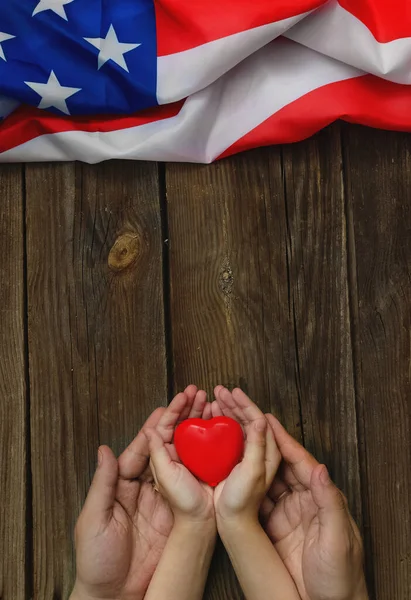 Flag Day. Memorial Day or 4th of July. Hands of father and child hold a heart next to the American flag. National patriotic holiday. Greeting card or banner. Layout. Vertically