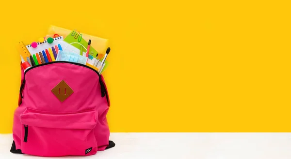 Back to school. Backpack for school with bright colorful office supplies and medical mask on yellow background. Stationery for school children's studies. Greeting card or banner for sale. Copyspace