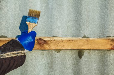 Elinde boya fırçası tutan, ahşap kaplama uygulayan mavi eldivenli bir adam. Marangoz elindeki fırçayla ahşap bir tahtayı boyuyor..
