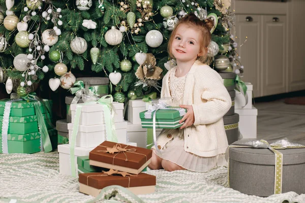 Merry Christmas and Happy Holidays! Beautiful little girl in a dress with a gift in her hand sitting near the Christmas tree. Winter holidays. Happy New Year. Portrait. Winter holidays concept