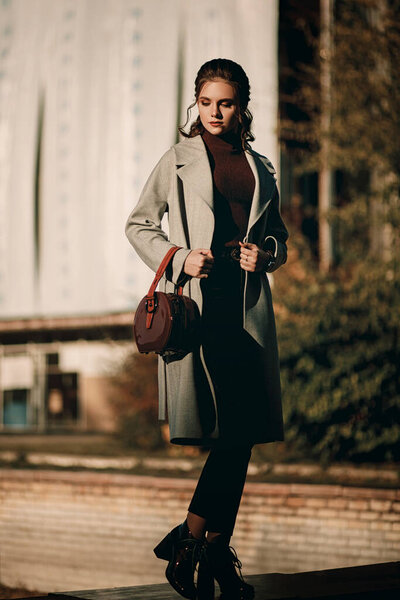 Young fashionable beautiful girl in a long gray coat, black jeans and with a dark red bag. Portrait shooting of a stylish girl. Facial shooting, street style and fashion. Professional model.