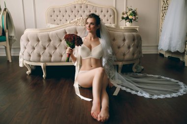 Gelinin sabahı. Güzel seksi sarışın kız beyaz dantel iç çamaşırlarıyla poz veriyor. Beyaz iç çamaşırlı güzel seksi bir kızın portresi. Beyaz çarşaflar içinde baştan çıkarıcı genç bir kadın ve bir peignoir. Bir buket kırmızı gülle güzel bir gelin. Yatak odası çatışması.