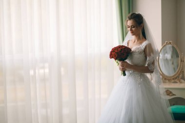 Beyaz ipek sabahlık, kıvırcık saç stili ve uzun peçeli güzel gelinin portresi yatak odasının penceresinin yanında duruyor. Gelinin sabah hazırlığı. Arka plan