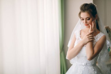 Beyaz ipek sabahlık, kıvırcık saç stili ve uzun peçeli güzel gelinin portresi yatak odasının penceresinin yanında duruyor. Gelinin sabah hazırlığı. Arka plan