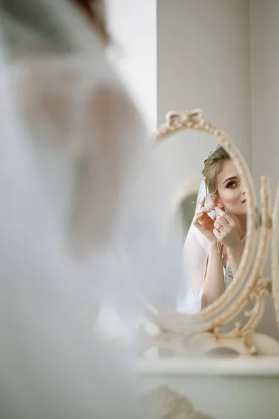 Portrait of a gorgeous bride looking in the mirror and putting on her wedding earrings. Beautiful bride putting on her wedding earrings in the room in front of a small white mirror.