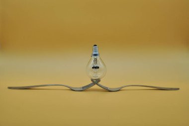 İki iç içe geçmiş çatal üzerinde dengelenen ampul turuncu nötr zemin üzerinde izole edilmiş, kavramsal fotoğraf elektriğin doğru bölünmesini gösterir.