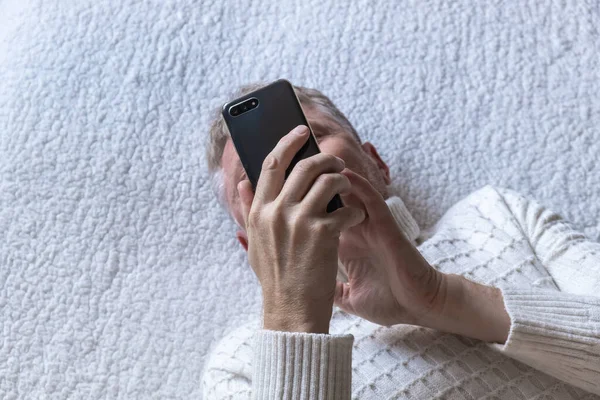 Orta yaşlı bir adam kanepeye uzanıp haberleri cep telefonundan izliyor. Mesajlaşıyorum, borsayı kontrol ediyorum. Seçici odaklanma. Yakın çekim. Üst görünüm