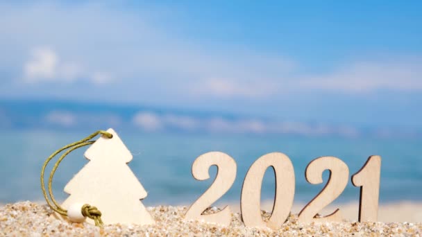 Números de madera 2021 y un puesto de decoración de árboles de Navidad en la arena de la playa. En el fondo, piernas borrosas por un hombre que pasa. — Vídeos de Stock