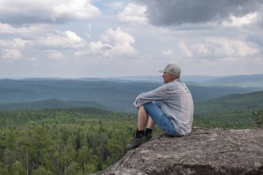 Özgür, orta yaşlı bir adam dağın tepesinde oturur ve dağ vadisinin güzel manzarasının tadını çıkarır. Seyahat konsepti. Yalnızlık, doğayla bütünlük. Boşluğu kopyala.