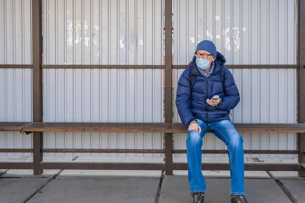 Tıbbi maskeli orta yaşlı bir adam sonbaharda toplu taşıma durağında oturur ve otobüs, tramvay ve tramvay bekler. Akıllı telefon kullanarak konum bilgisini takip eder ve uzaklığa bakar..