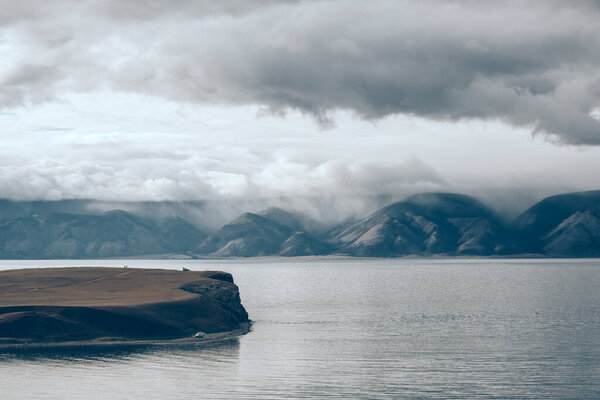 Драматические облака над озером и в горах. Серое туманное утро, облачная погода. Низкие облака создают мрачный вид на далекие холмы и горы. Озеро Байкал, Россия.