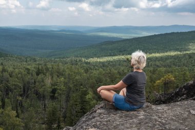 Orta yaşlı bir kadın, ormanlık bir tepe vadisinin çarpıcı manzarasıyla bir dağın tepesinde yoga ve meditasyon yapıyor..