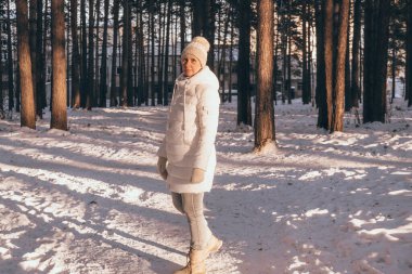 Sıcak ceketli, şapkalı ve eldivenli mutlu orta yaşlı kadın gün batımında karlı bir kış parkında yürüyor. Kış tatili.