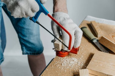 İş eldivenli bir adam küçük bir tahta parçasını testereyle kesiyor. Ev ve bahçe için el yapımı raf üretimi için malzeme hazırlama. ÇEVİR.