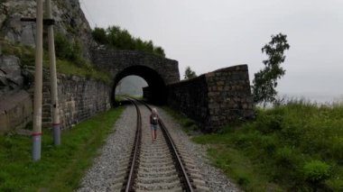 4K İHA, tünelden geçen tren raylarında yürürken sırt çantalı bir kadın yolcuyu takip ediyor. Yerel seyahat ve hafta sonu yürüyüşleri. Sisli bulutlu bir yaz günü, yürüyüş için harika bir gün..