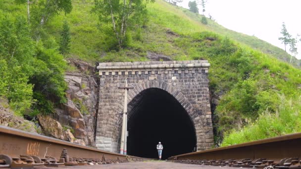 バックパック付きの男性観光客がトンネルを通って敷設鉄道のレールに沿って歩いています スローモーション 地元の旅行 新しい場所を探索し 週末にハイキング アクティブレクリエーション 観光の概念 — ストック動画