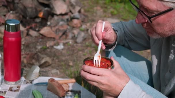 野餐的性质 一位中年男子戴着眼镜 津津有味地吃着番茄酱罐头鱼 背景是一张附有蔬菜和面包的临时桌子 好吃的小吃 — 图库视频影像