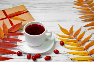 Sonbahar yaprakları ve bir hediye ile ahşap bir zemin üzerinde gül kalçalı beyaz çay fincanı. Sonbahar yaşam tarzı