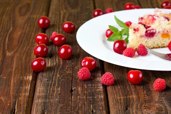 Several raspberries and cherries around white plate with cake — Stock Photo, Image