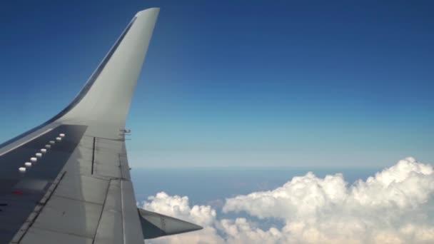 白い雲の切れ間から青空の飛行機が飛んでいます。窓から飛行機の翼のビュー。旅行と交通機関の概念. — ストック動画