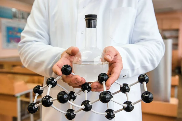 Molekül modeli öğretmenin ellerinde. Beyaz önlüklü bir bilim adamı elinde bir matara ve bir molekül modeli tutuyor. Mataralı matara. Atomik yapı.
