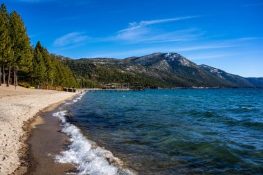 Gündüz vakti Tahoe Gölü sahilinde. Dalgalar kuma çarpar. Arka planda dağların zirveleri kar.. 